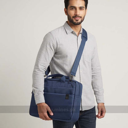 A man wearing a light gray long-sleeve shirt and dark jeans carries a navy blue crossbody laptop bag over his shoulder against a plain light gray studio background.