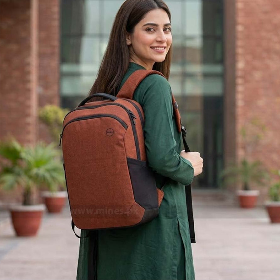 A woman wearing a dark green dress is shown from the back, carrying a rust-brown and black laptop backpack in an outdoor courtyard setting with brick buildings and potted plants.